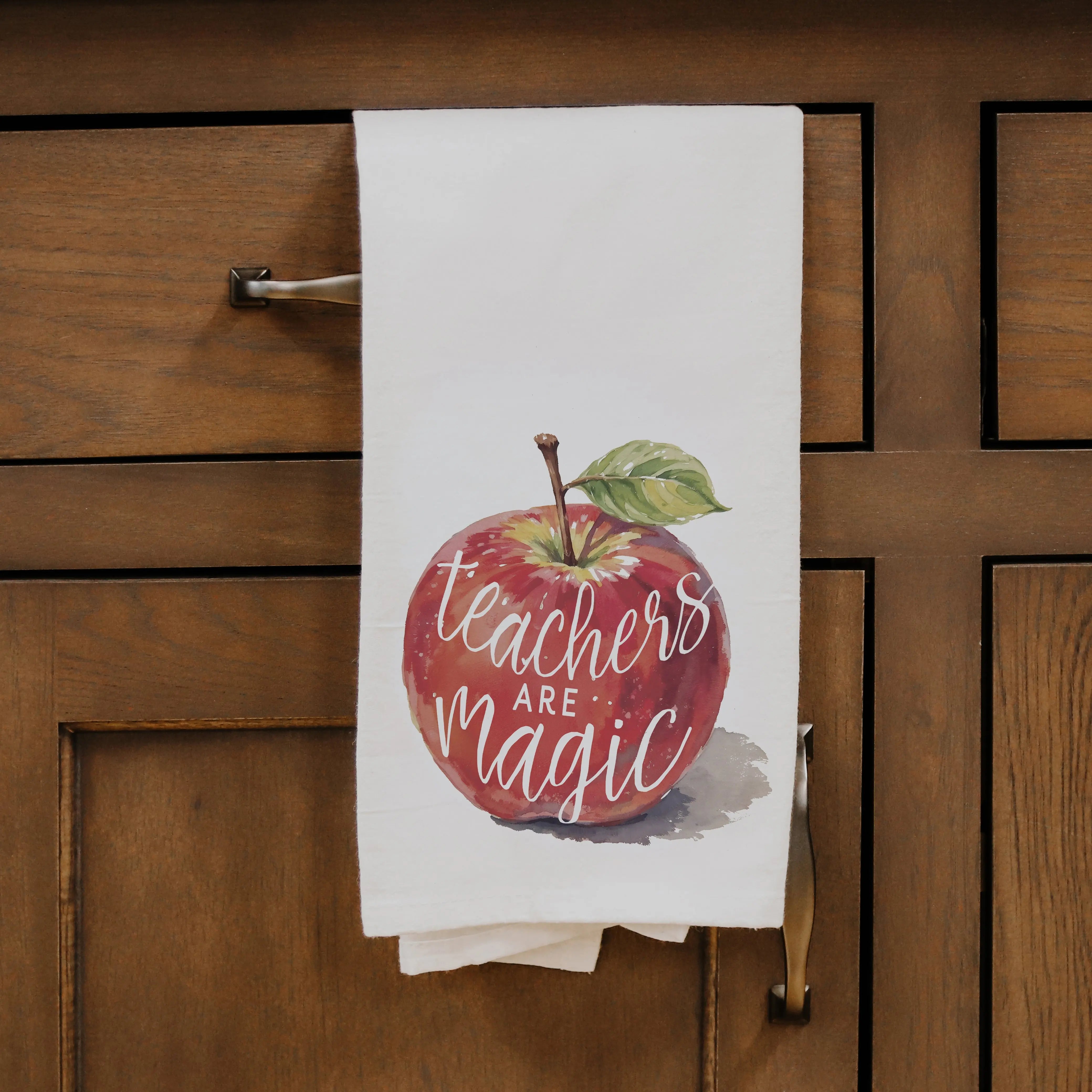 Tea towel with an apple design and 'Teachers are Magic' text hanging on a wooden cabinet.