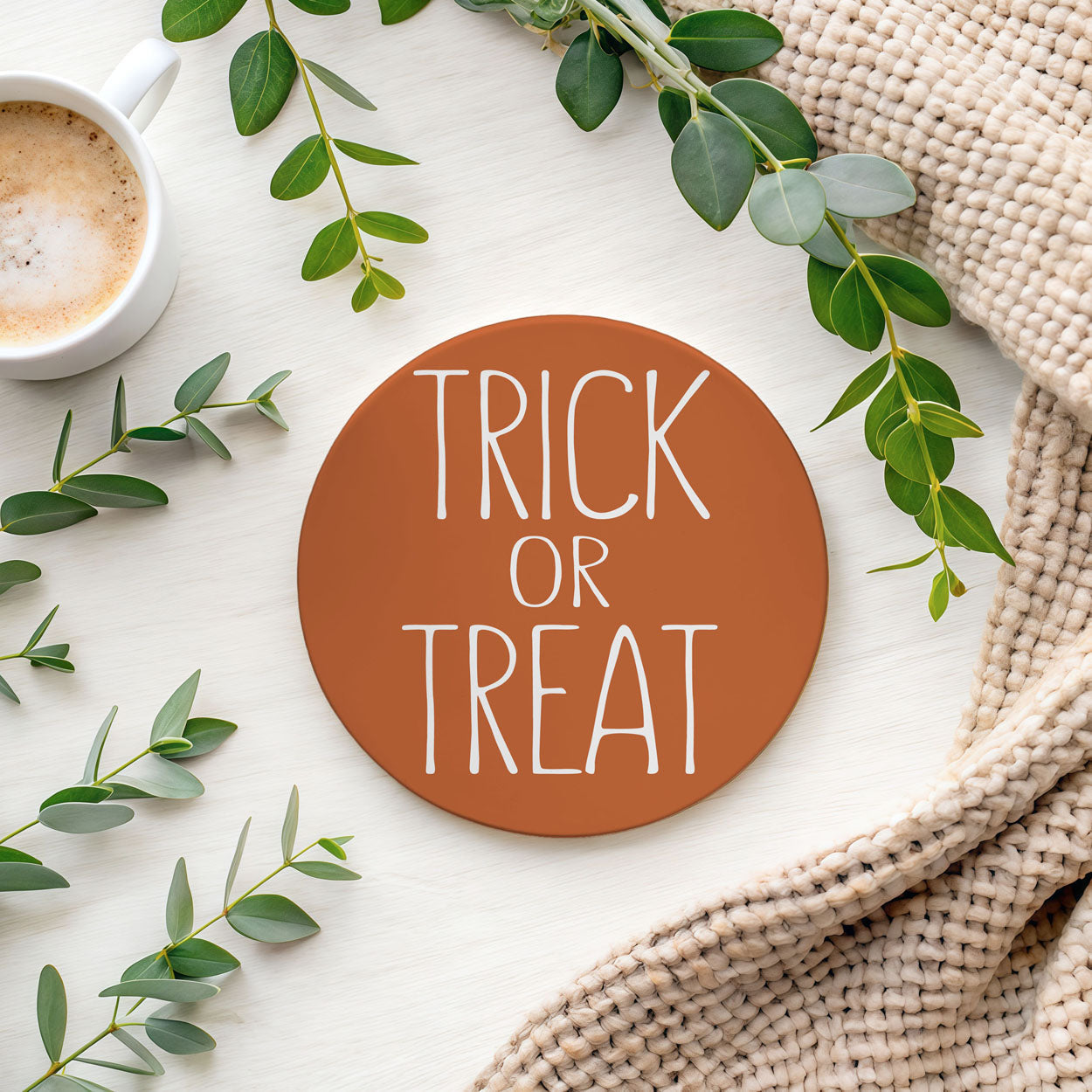 Round brown coaster with 'TRICK OR TREAT' text on a white surface with green leaves and a cup of coffee.