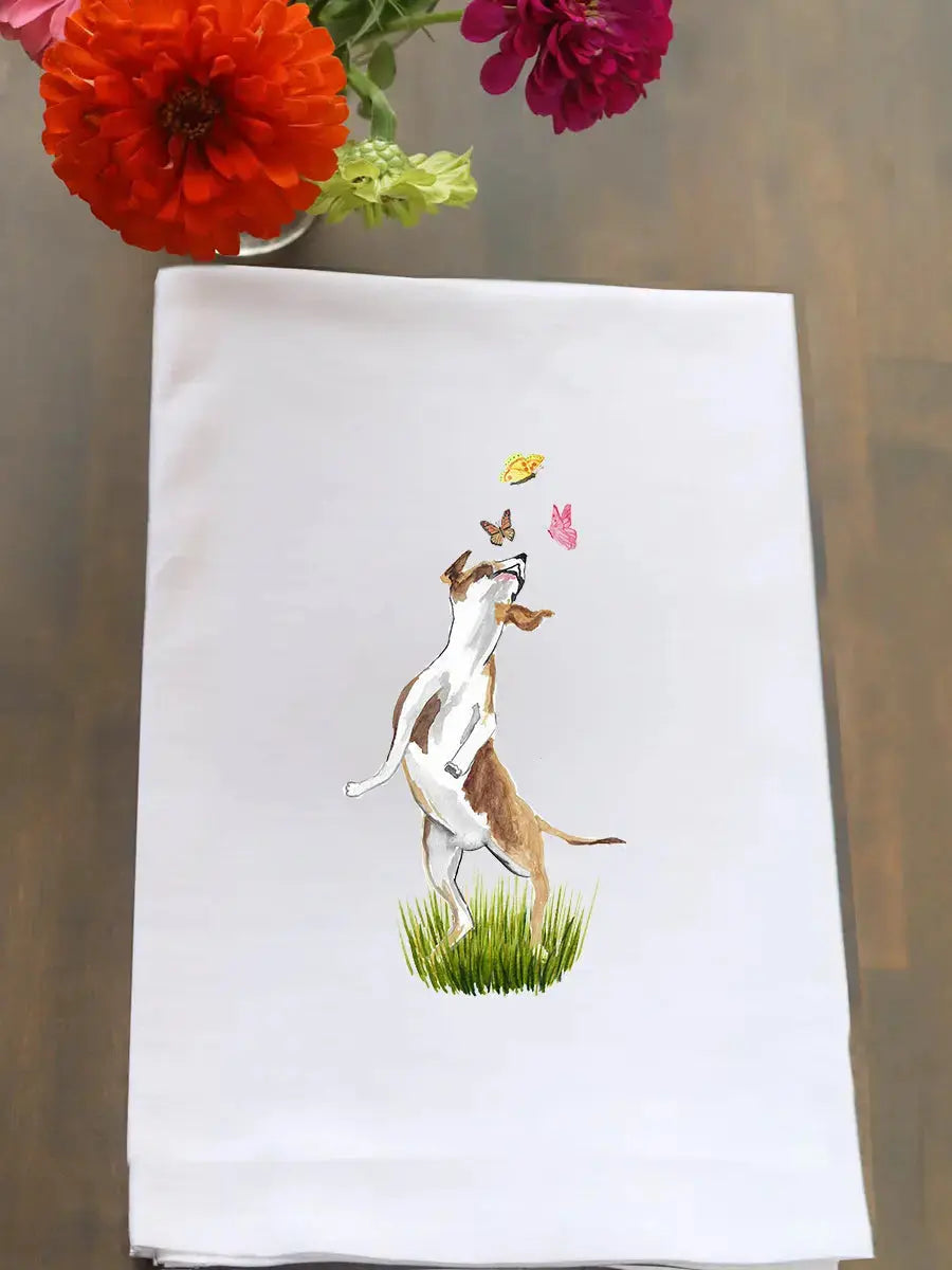 A white kitchen towel featuring a watercolor illustration of a dog playing with butterflies on grass, displayed on a wooden table with flowers in the top corner.