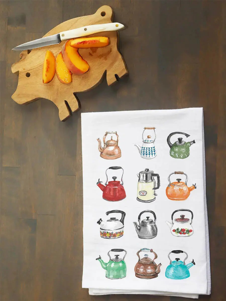 A white kitchen tea towel printed with a grid of colorful illustrated kettles, laid on a dark wooden table next to a wooden peacock-shaped cutting board with a knife and peach slices.