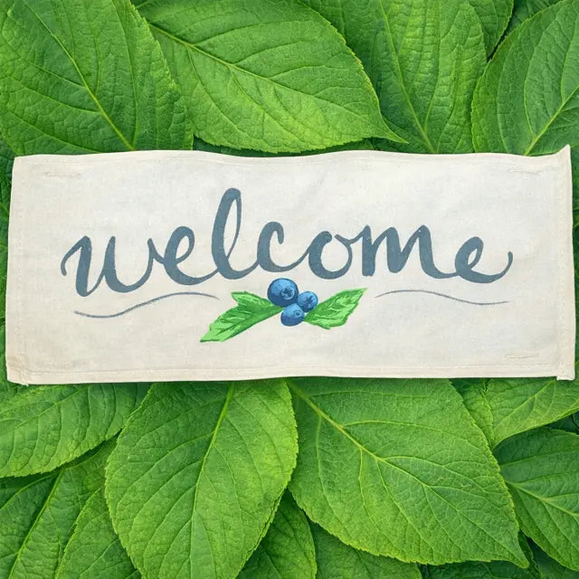 A rectangular cream-colored fabric sign with the word 'welcome' in dark blue script, decorated with a small green leaf and blue berry illustration, laid flat on a bed of vibrant green leaves.