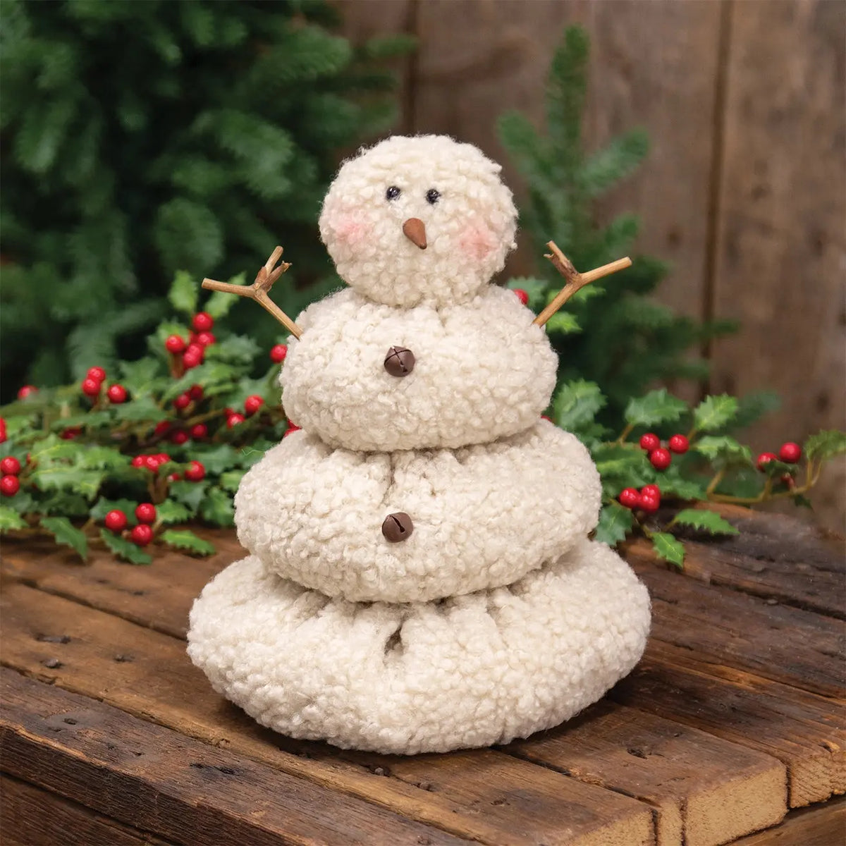 A small handmade snowman decoration made from three stacked cream bouclé fabric discs, with twig arms, a brown nose, black embroidered eyes, pink cheeks, and two brown buttons. It is displayed on a rustic wooden surface with green pine branches and red berries in the background.