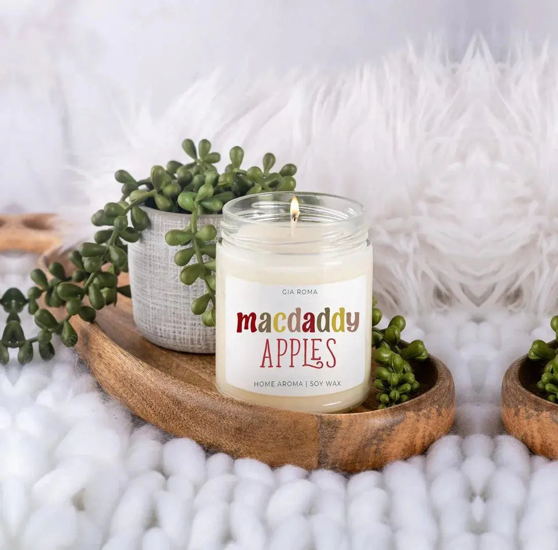 Candle labeled 'macdaddy APPLES' on a wooden tray with decorative plants.