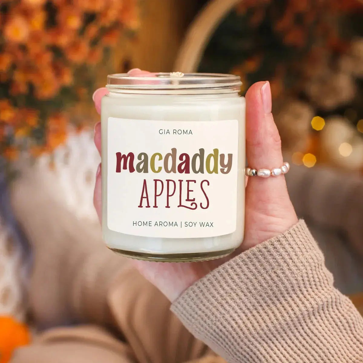 Hand holding a candle labeled 'macdaddy APPLES' with a blurred autumn background