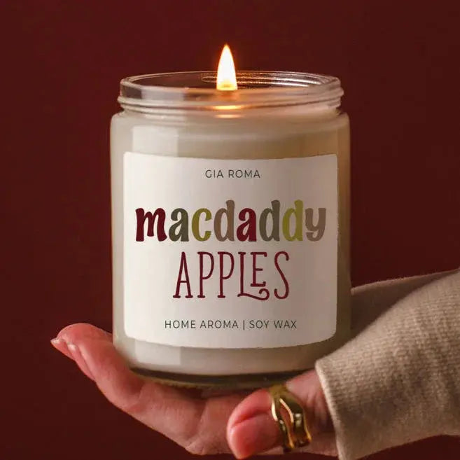 Candle labeled 'macdaddy APPLES' held by a hand with a gold ring against a dark background