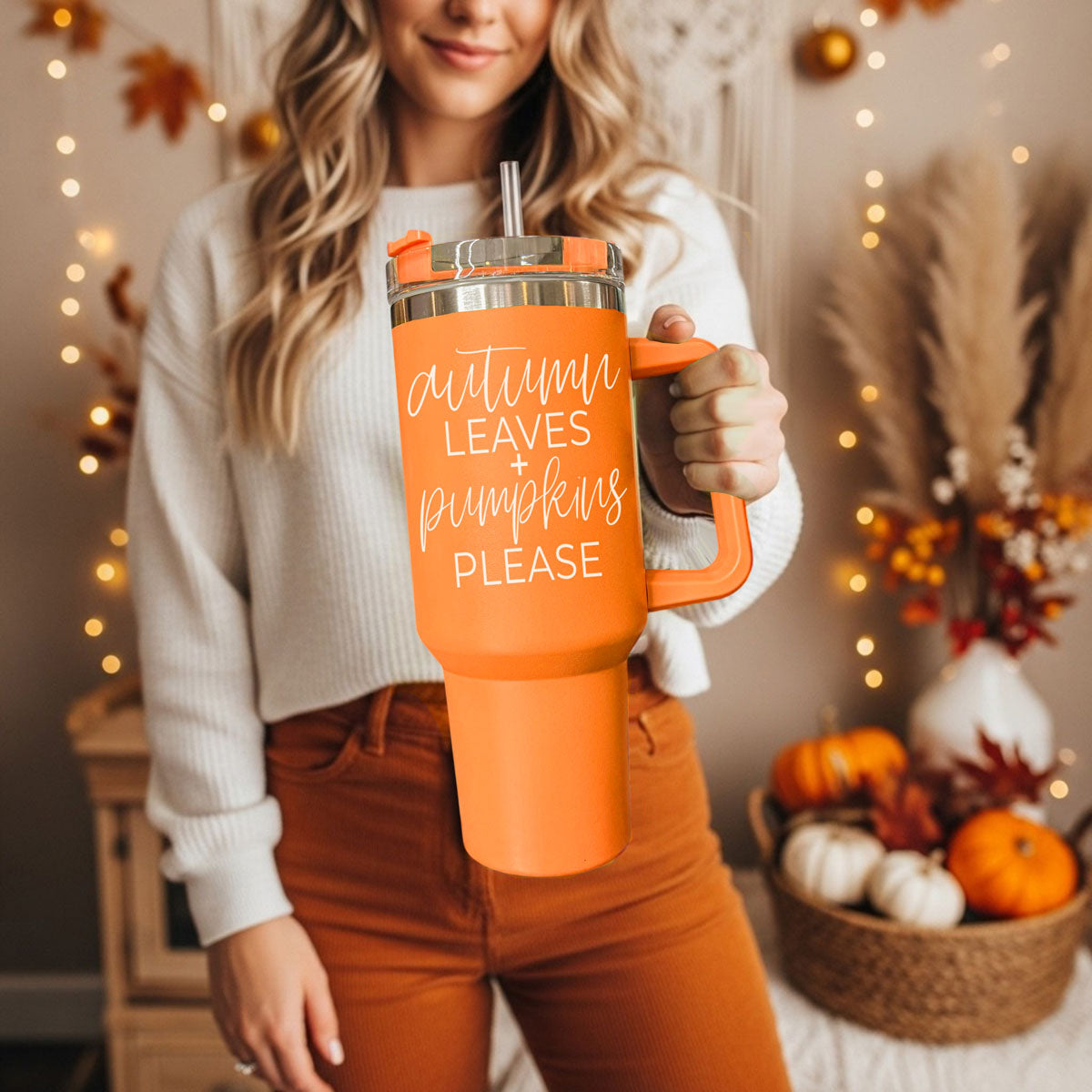 Person holding an orange mug with text in a cozy indoor setting with pumpkins and lights.