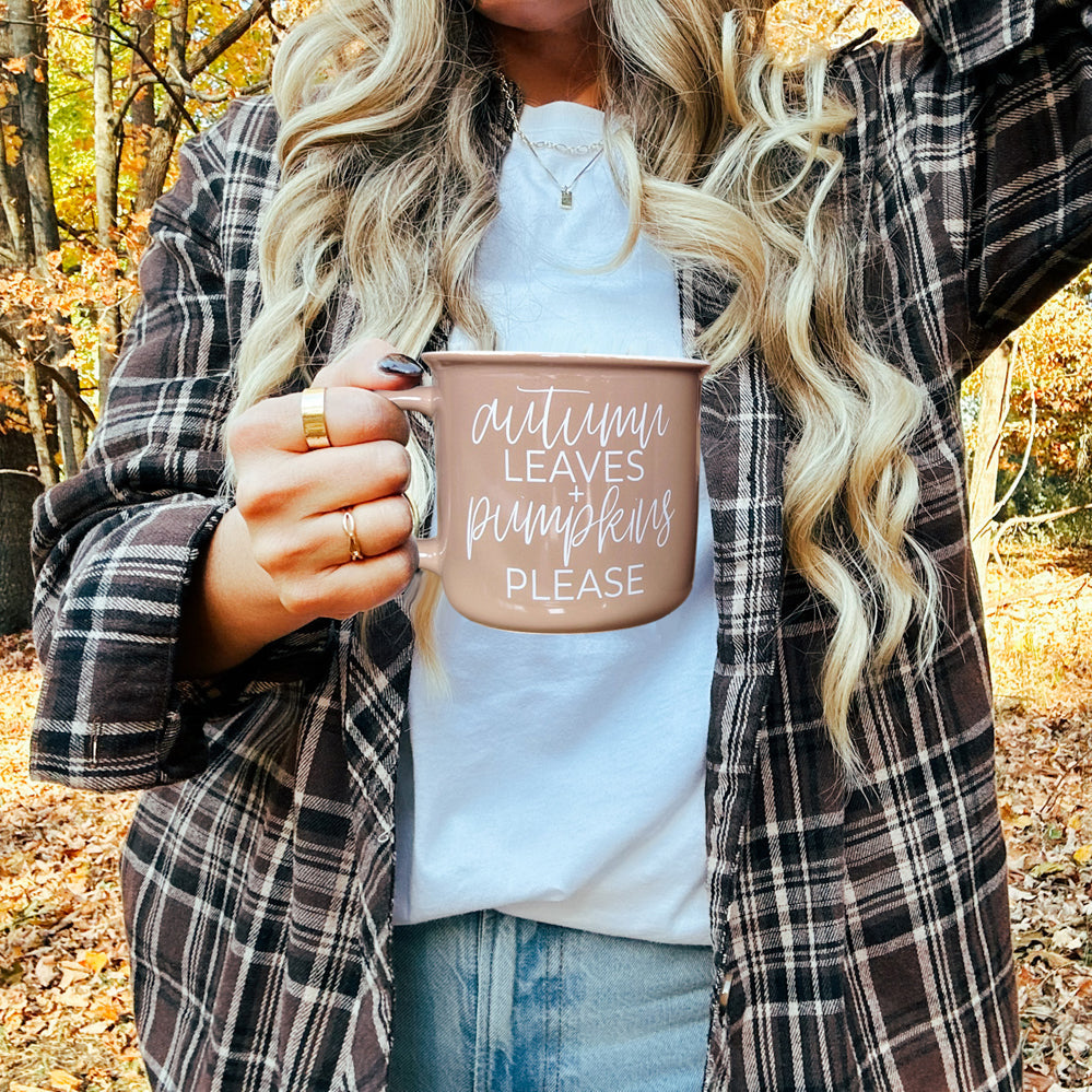 Person holding a mug with 'Autumn leaves, pumpkins, please' text in an outdoor setting with fall foliage.