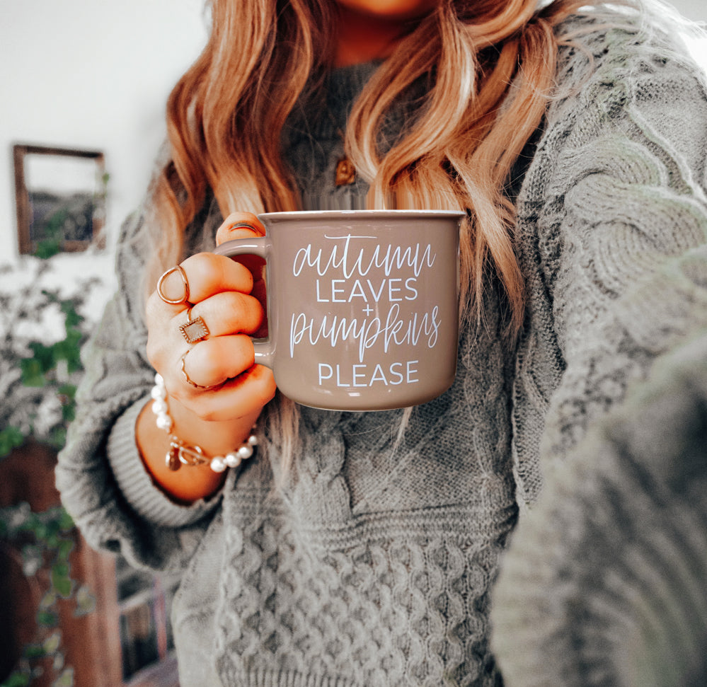 Person holding a mug with 'Autumn leaves & pumpkins please' text, wearing a cozy sweater indoors.