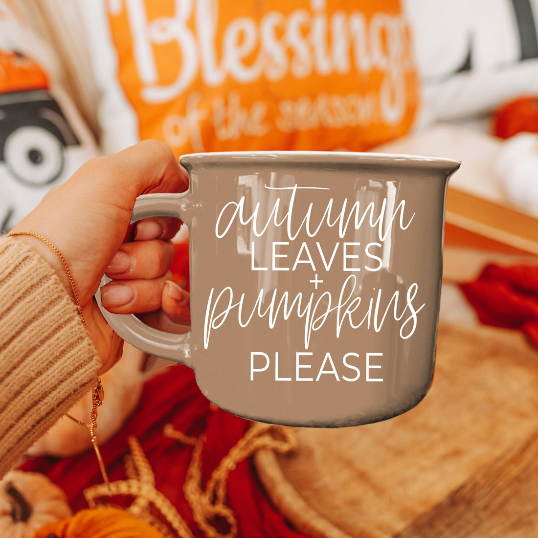 Hand holding a mug with 'Autumn leaves + pumpkins please' text, blurred background with autumn decorations.