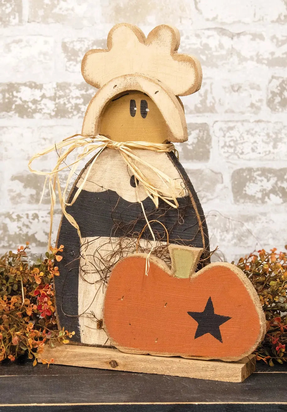 A rustic wooden decorative figure with a cowboy hat, painted black and beige body, and a large orange pumpkin in front with a black star, set against a distressed white brick wall and autumn foliage.