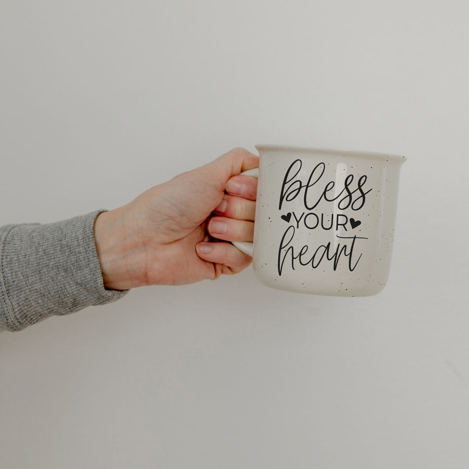 Hand holding a mug with 'bless your heart' text against a plain background