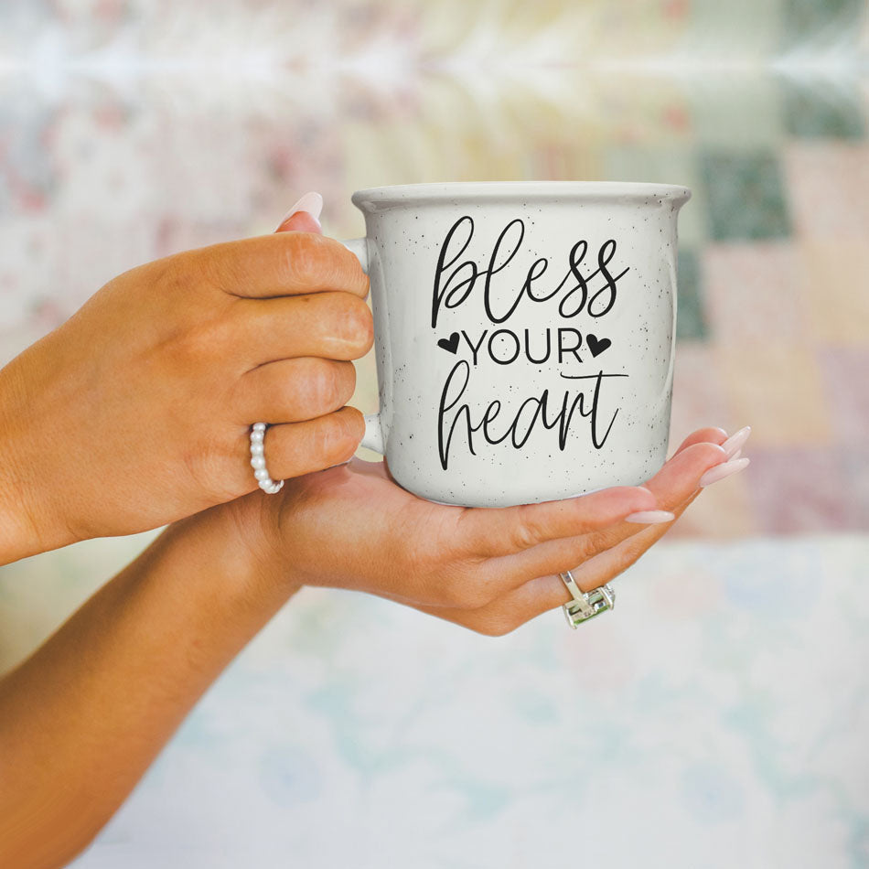 Hand holding a mug with 'bless your heart' text against a blurred background