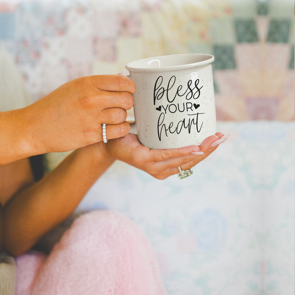Person holding a mug with 'bless your heart' text in a soft-focus indoor setting