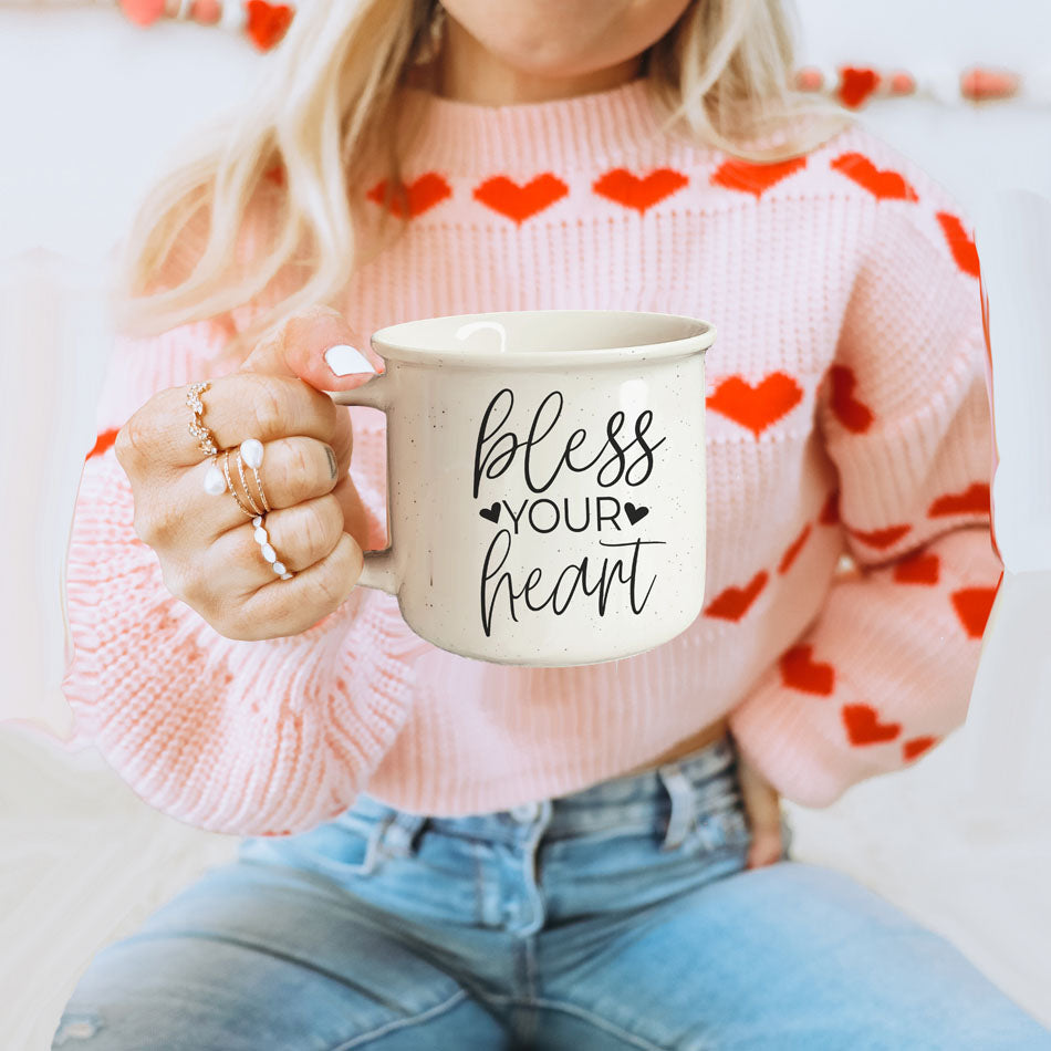 Person wearing a pink sweater with heart patterns, holding a mug with 'bless your heart' text.