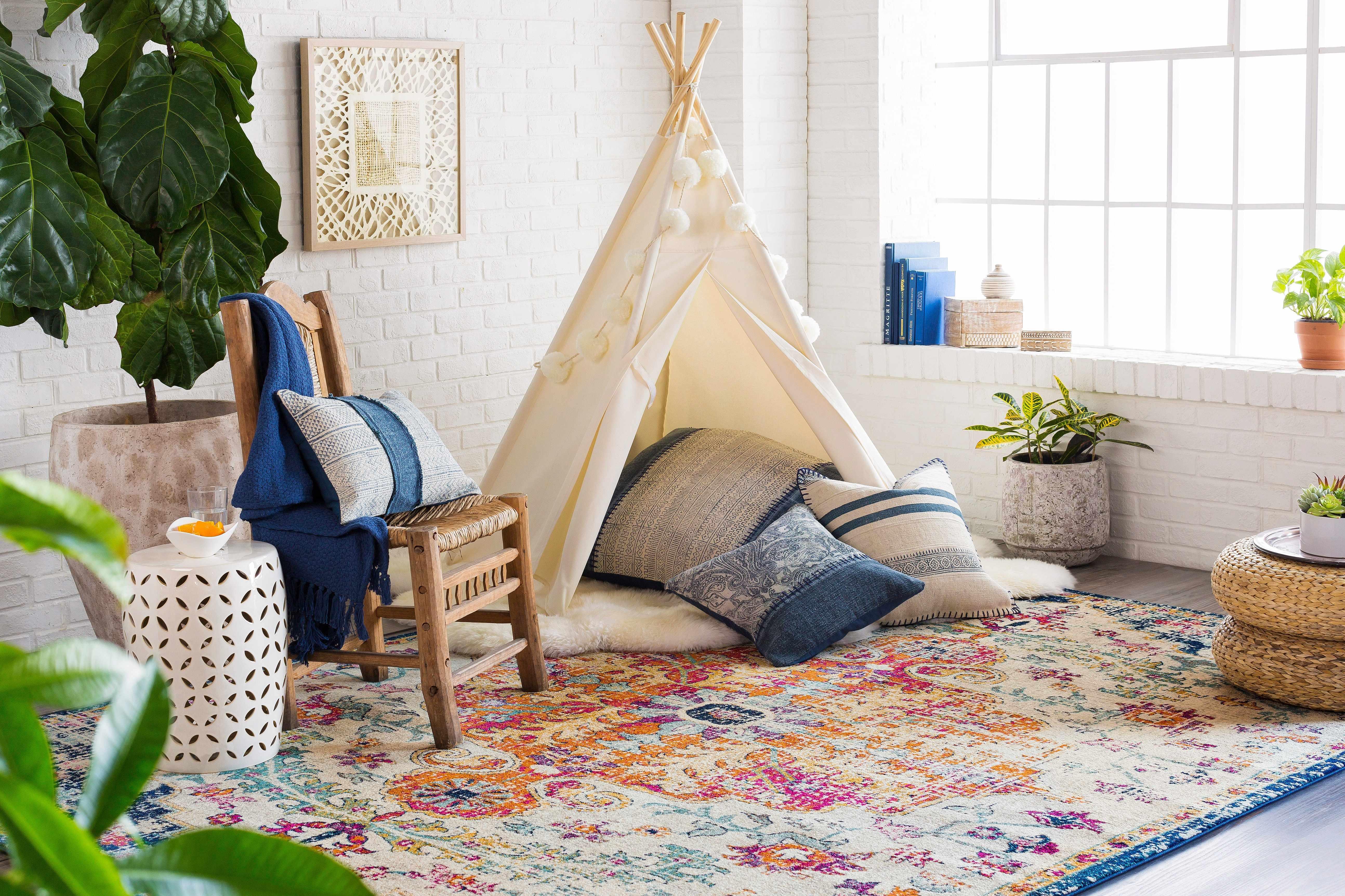 Cozy farmhouse living room with teepee, patterned rug, wicker chair, blue throw, and potted plants on white brick wall.