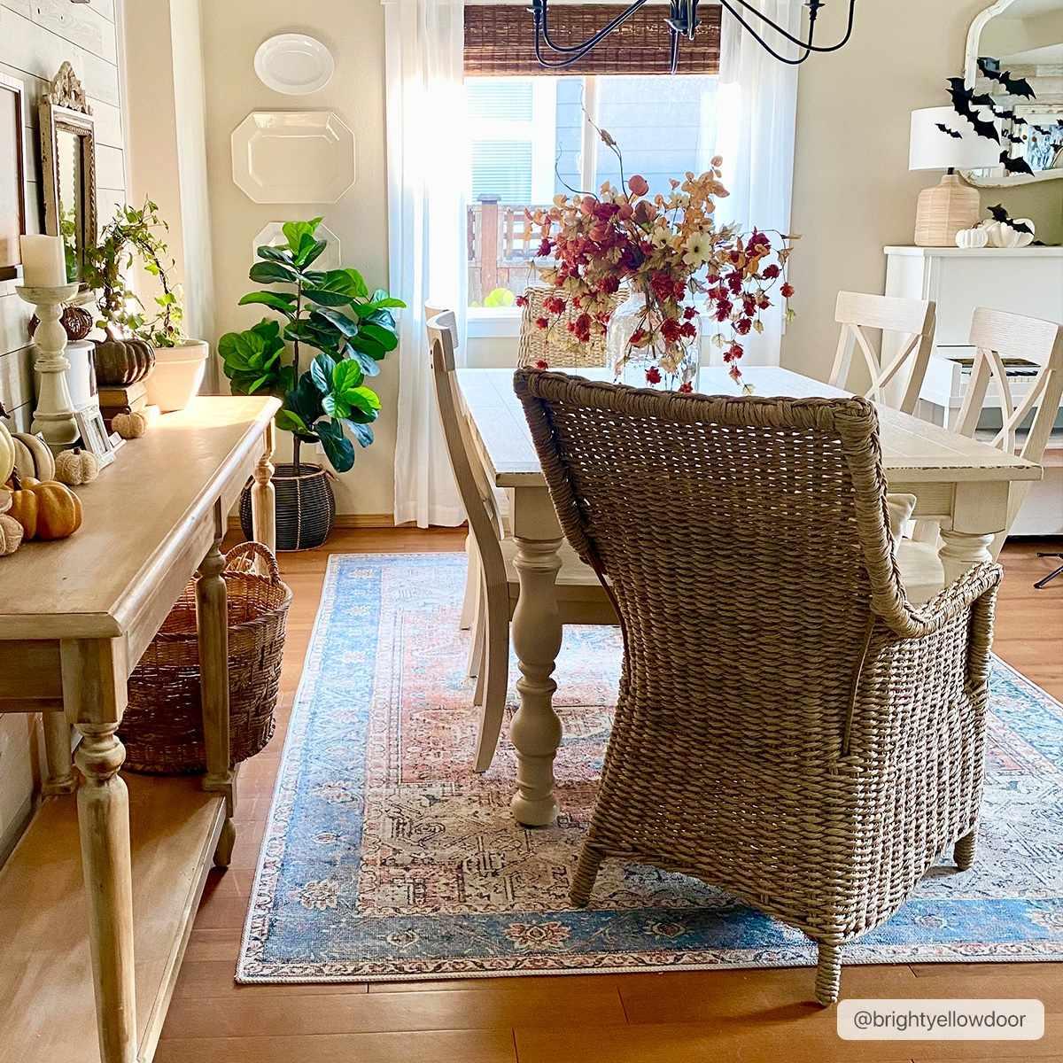 Cozy farmhouse dining room with wicker chair, rustic table, fall decor, and blue patterned rug