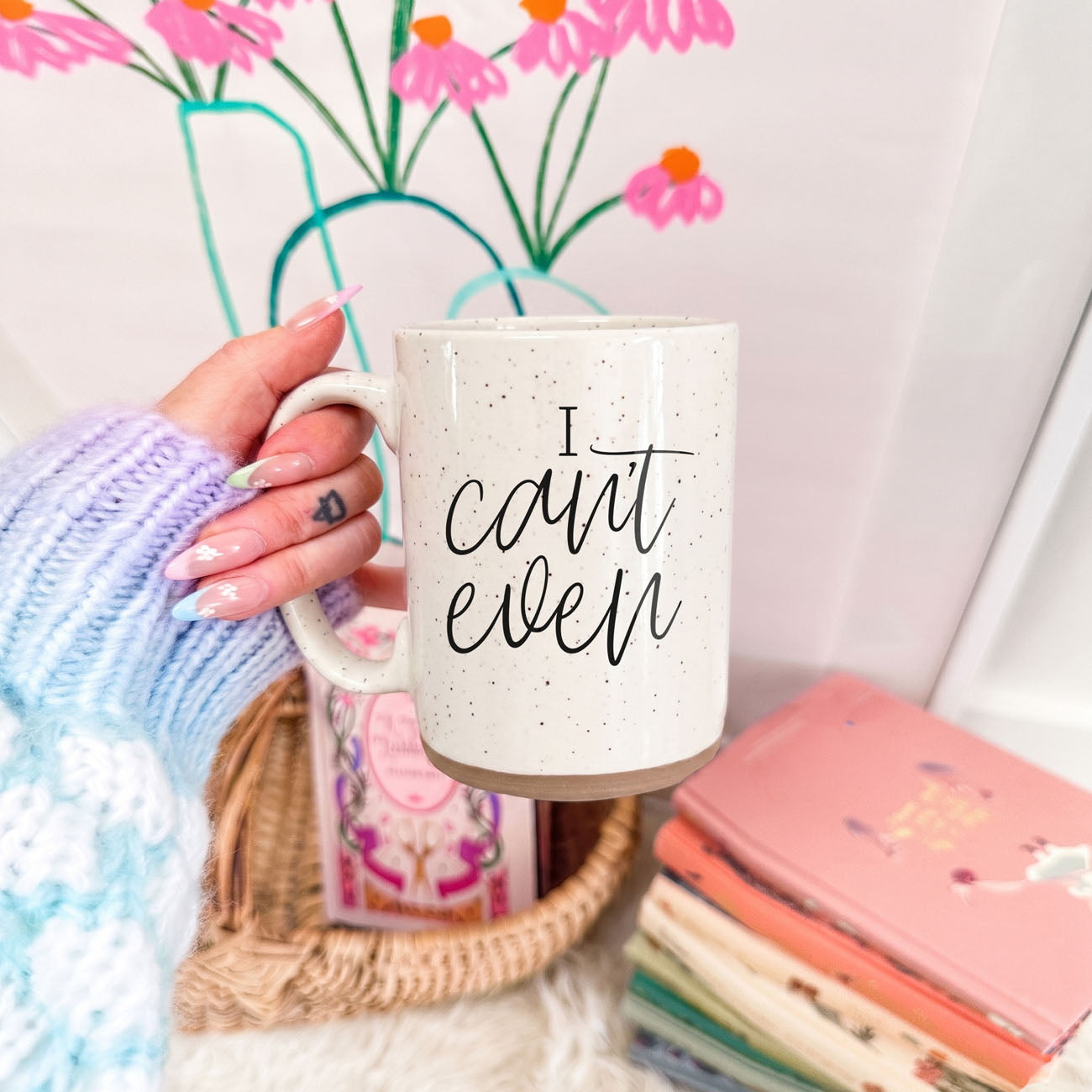 Person holding a mug with 'I can't even' text, surrounded by books and flowers.