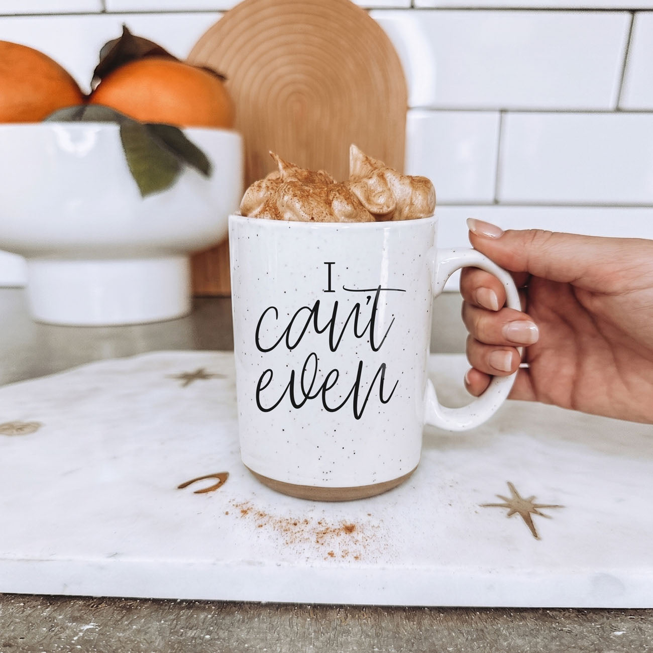 Hand holding a white mug with 'I can't even' text, filled with a hot beverage and whipped cream, on a kitchen counter.