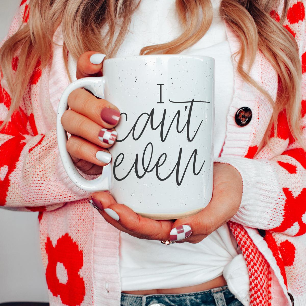 Person holding a white mug with 'I can't even' text, wearing a red and white patterned cardigan.