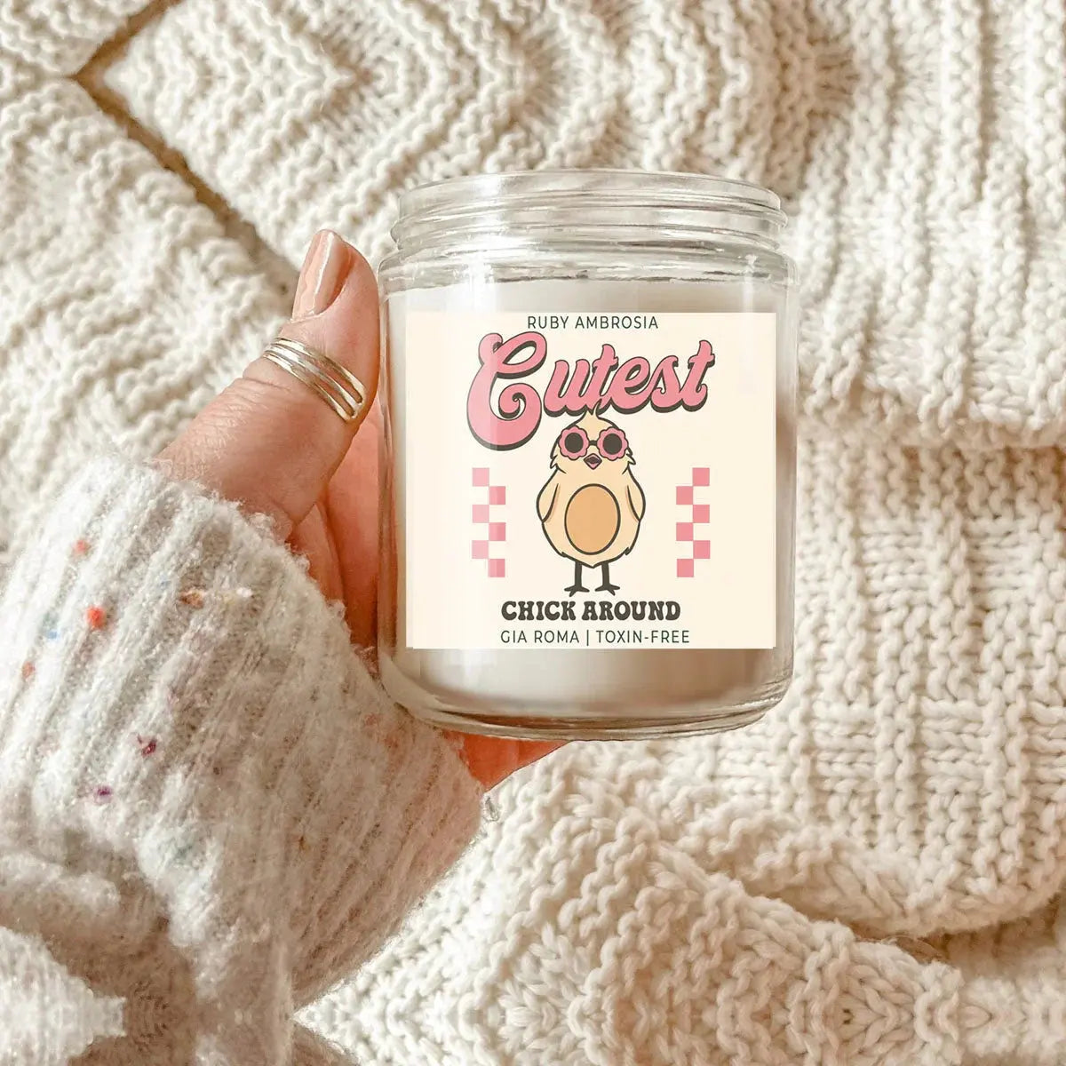 Hand holding a candle labeled 'Cutest Chick Around' against a textured beige background