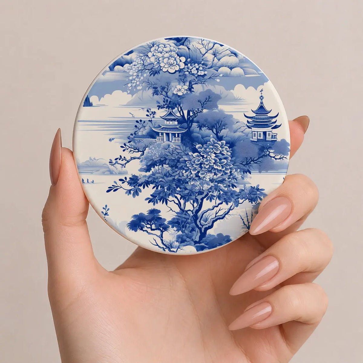 Hand holding a round compact mirror with blue floral and architectural design on a beige background