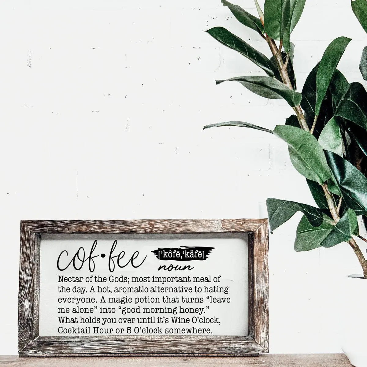 Decorative sign with coffee definition text in a wooden frame on a white wall with a plant.