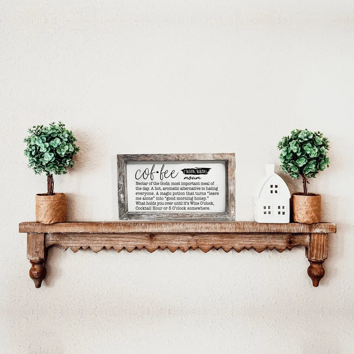 Wooden shelf with decorative items including a framed sign, potted plants, and a small house-shaped object on a light wall.