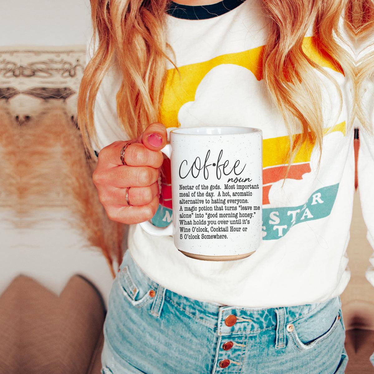 Person holding a mug with text, wearing a colorful shirt and jeans.