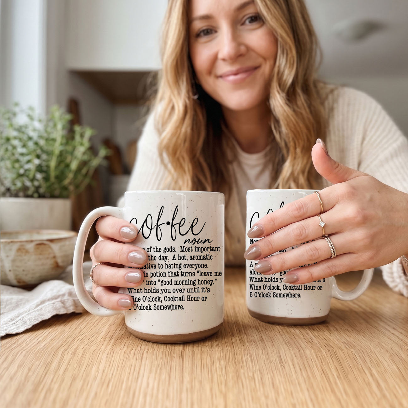 Woman holding two mugs with text design on a wooden table.