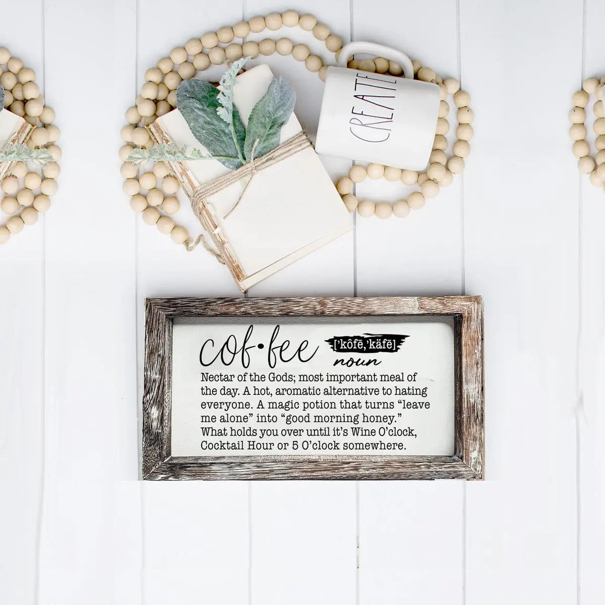 Decorative setup with a framed definition of 'coffee', mug, notebook, and beads on a white wooden surface.