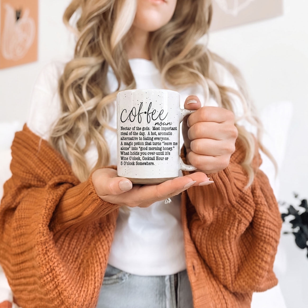 Person holding a mug with text, wearing an orange sweater and white shirt.