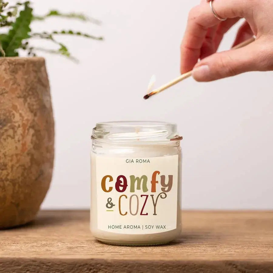Hand lighting a candle labeled 'Comfy & Cozy' on a wooden surface with a plant in the background.