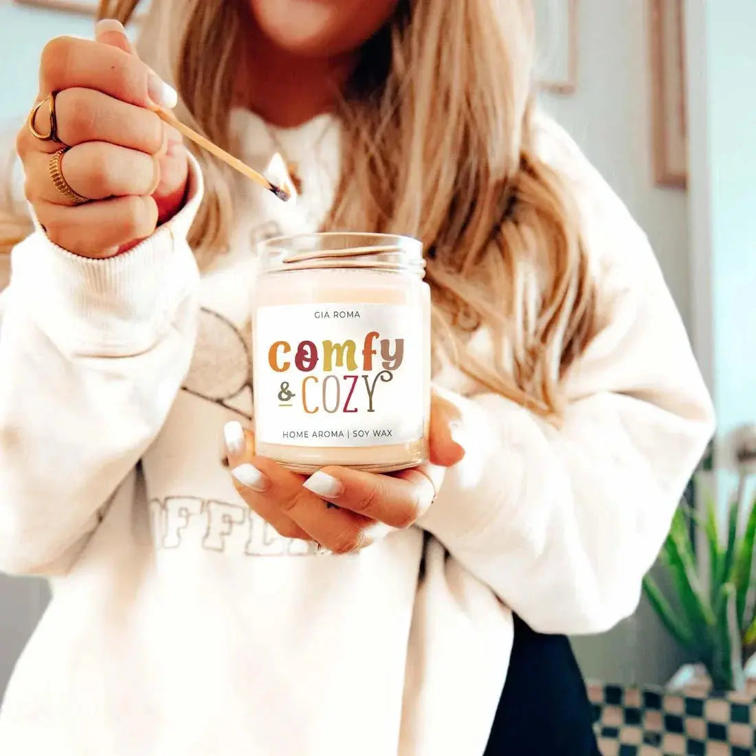 Person lighting a 'comfy & cozy' candle with a match.