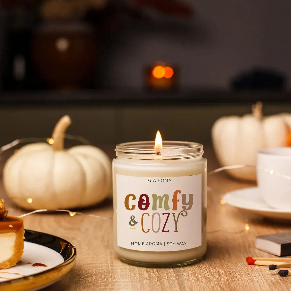 Candle labeled 'comfy & cozy' on a table with pumpkins and a cup in the background