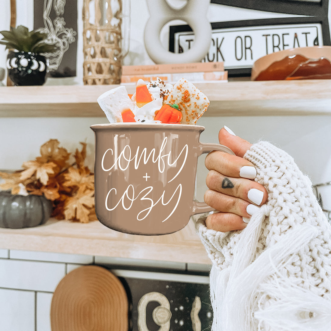 Person holding a 'comfy + cozy' mug with Halloween-themed treats on a shelf.
