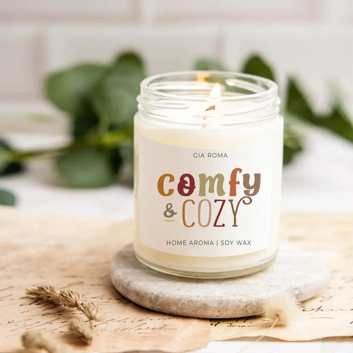 Candle in a jar labeled 'comfy & cozy' on a wooden surface with greenery in the background