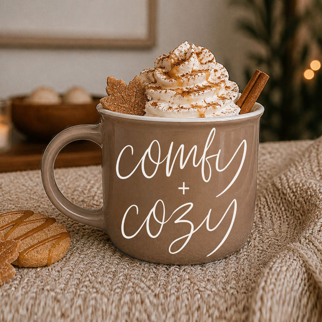 Mug with 'comfy + cozy' text, topped with whipped cream and a cinnamon stick, on a textured surface.