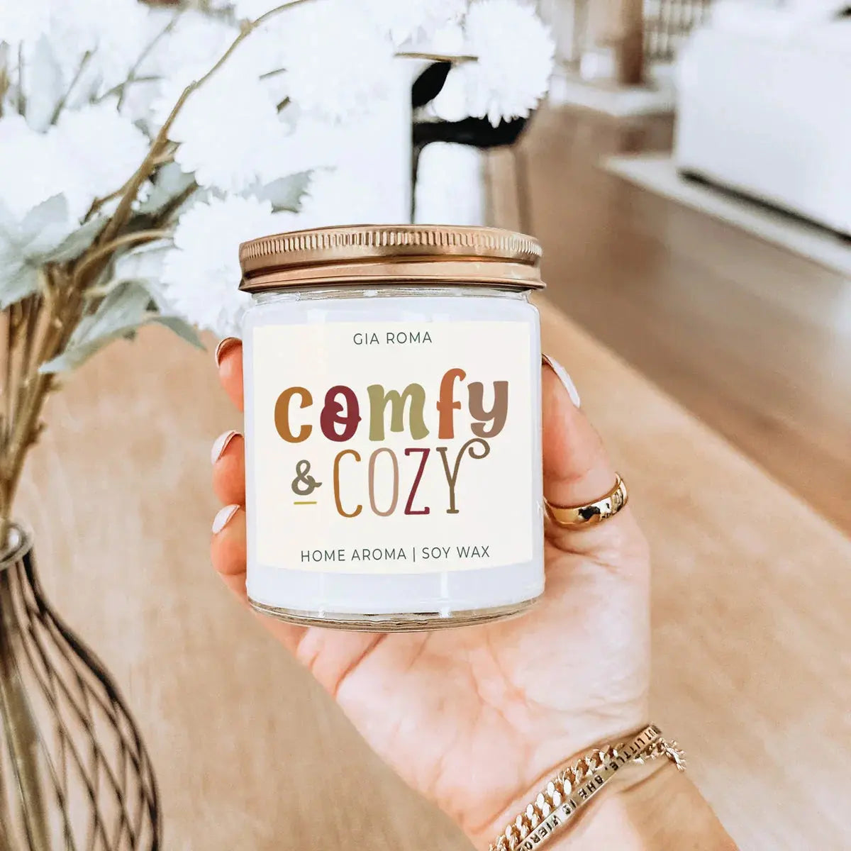 Hand holding a candle labeled 'comfy & cozy' with decorative elements in the background