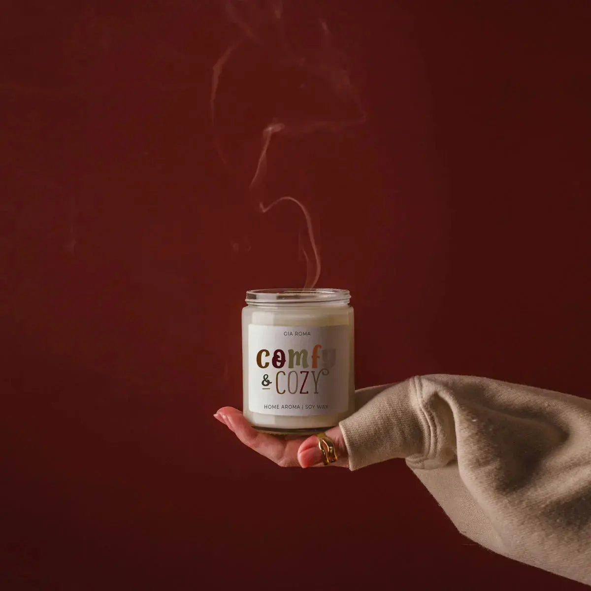 Hand holding a 'Comfy & Cozy' candle against a dark red background