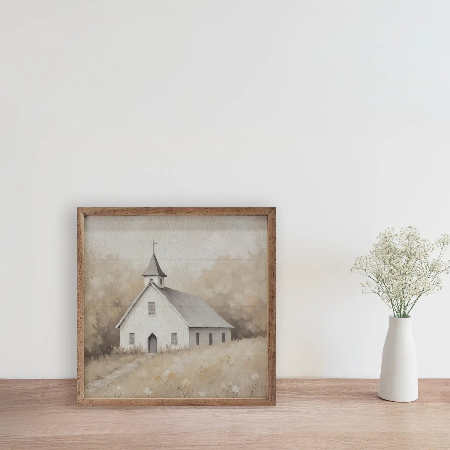 Framed artwork of a small white church on a wooden surface with a vase of flowers.