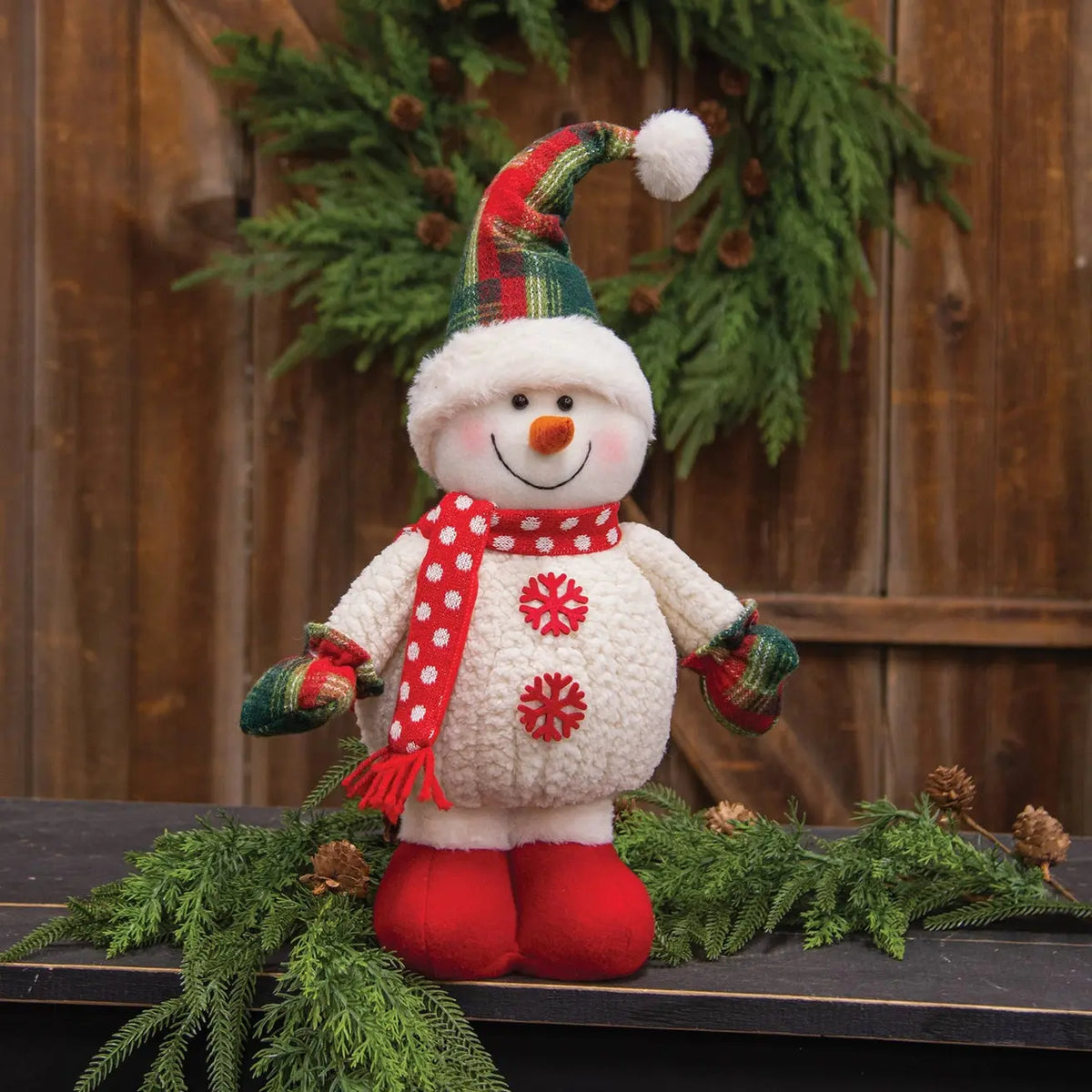 A crocheted snowman decoration standing on a dark wooden surface with greenery and a wooden fence backdrop. The snowman has a white textured body, red scarf with white polka dots, red mittens, red boots, and a red-and-green plaid hat with a white pom-pom.