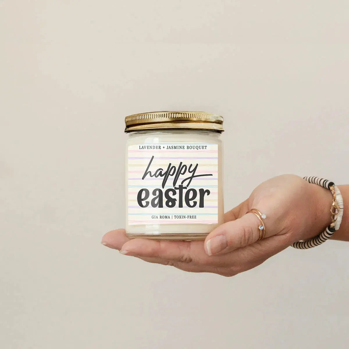 Hand holding a 'Happy Easter' scented candle with a gold lid on a beige background