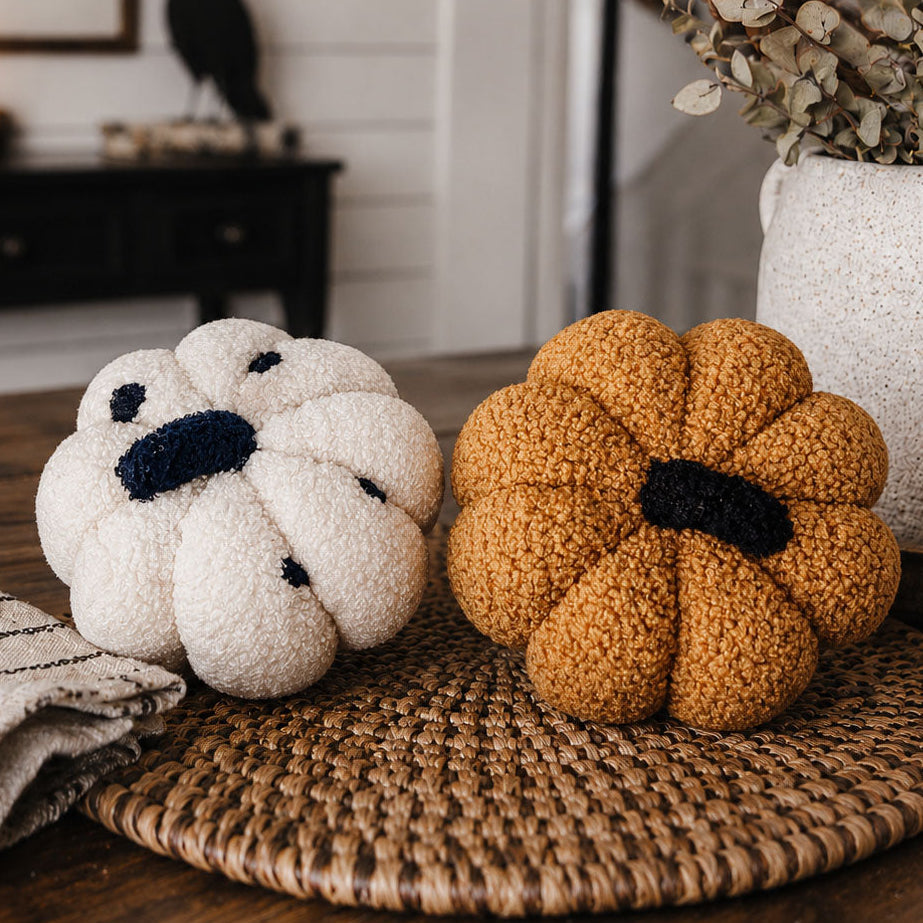 Two knitted pumpkins, one white and one brown, on a woven surface with a blurred indoor background.