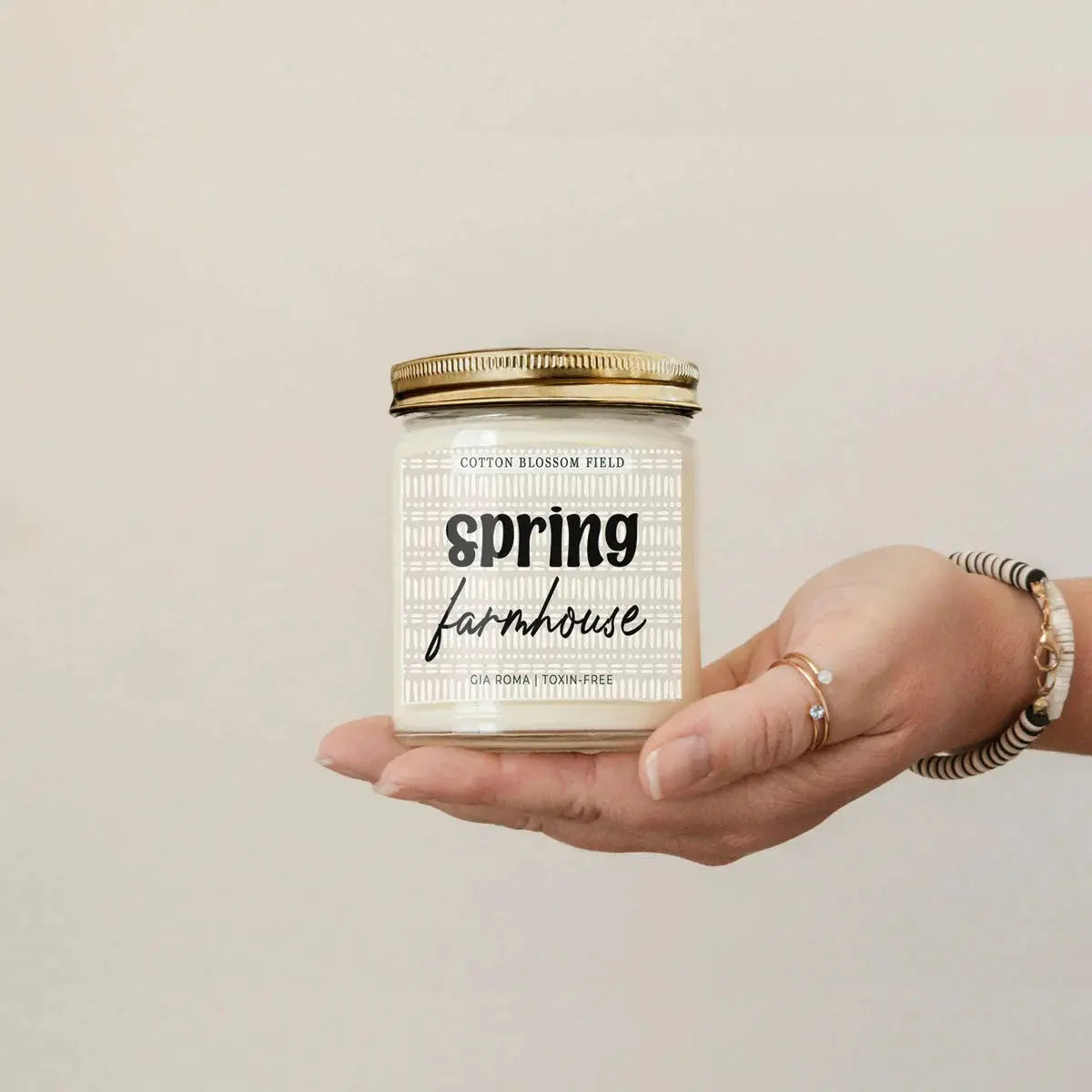 Hand holding a candle labeled 'Spring Farmhouse' against a beige background