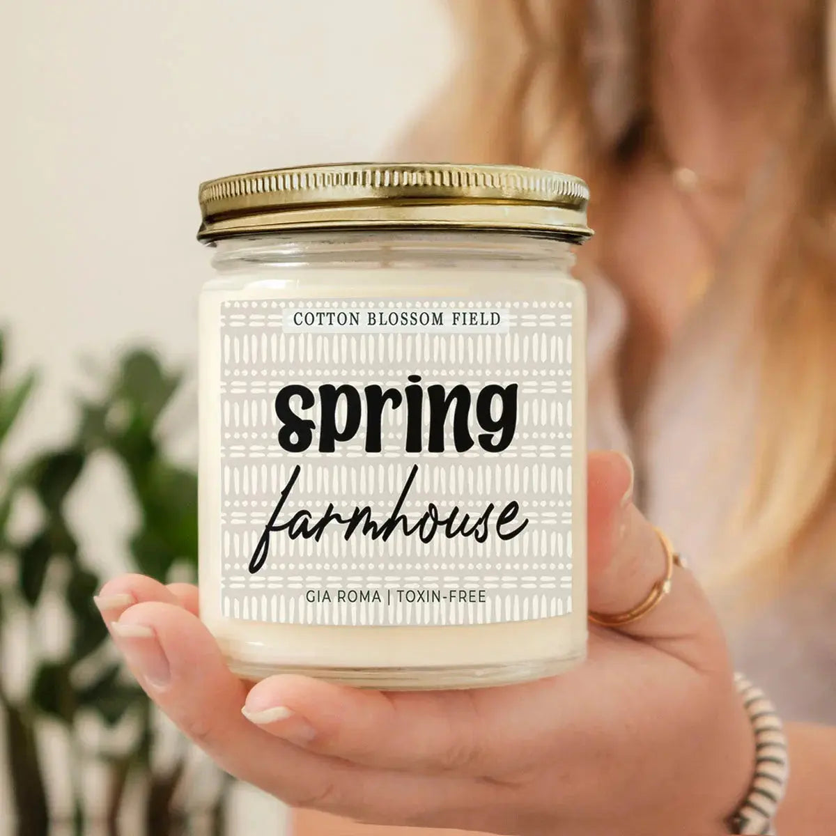 Hand holding a candle labeled 'Spring Farmhouse' with a blurred background