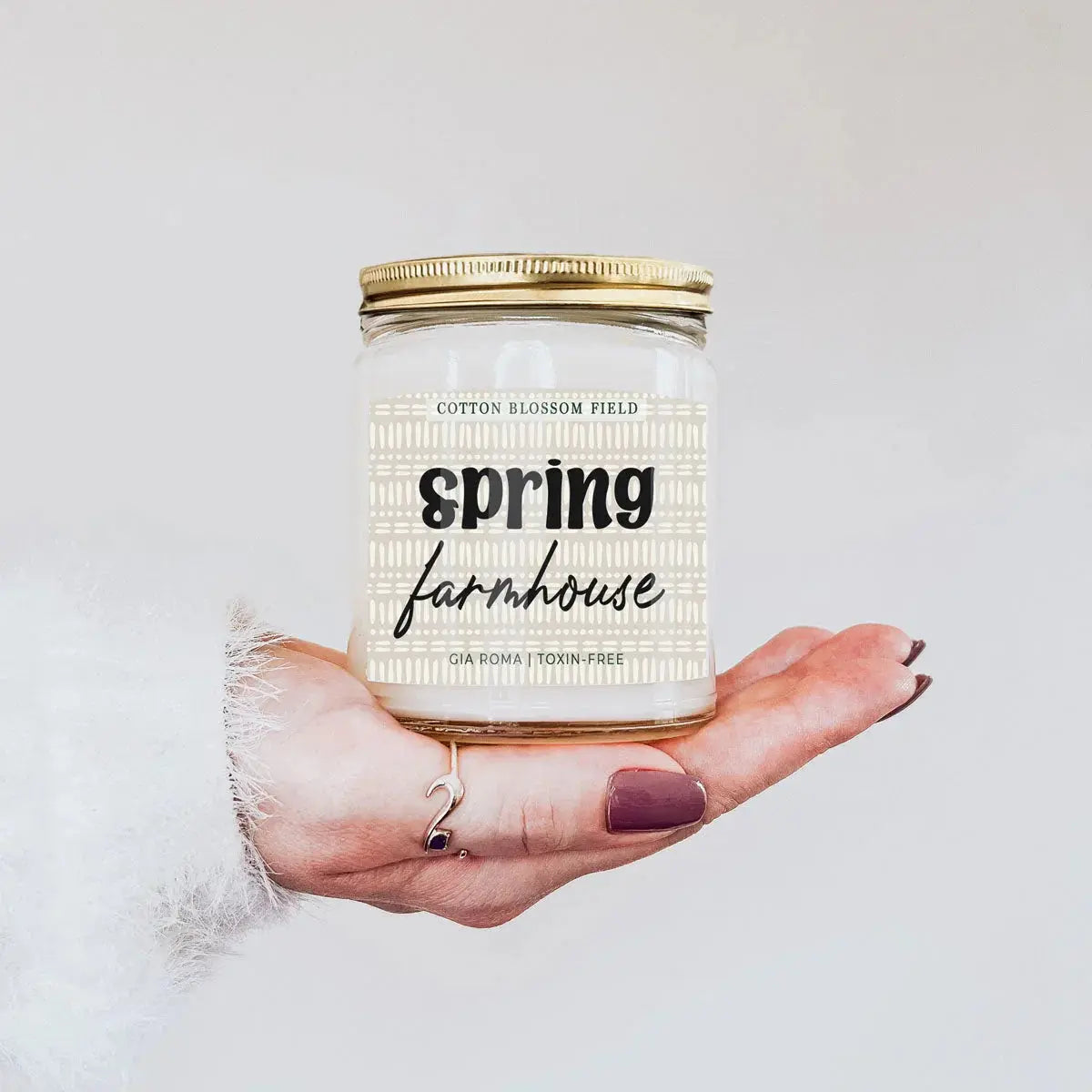 Hand holding a candle labeled 'Spring Farmhouse' against a light background