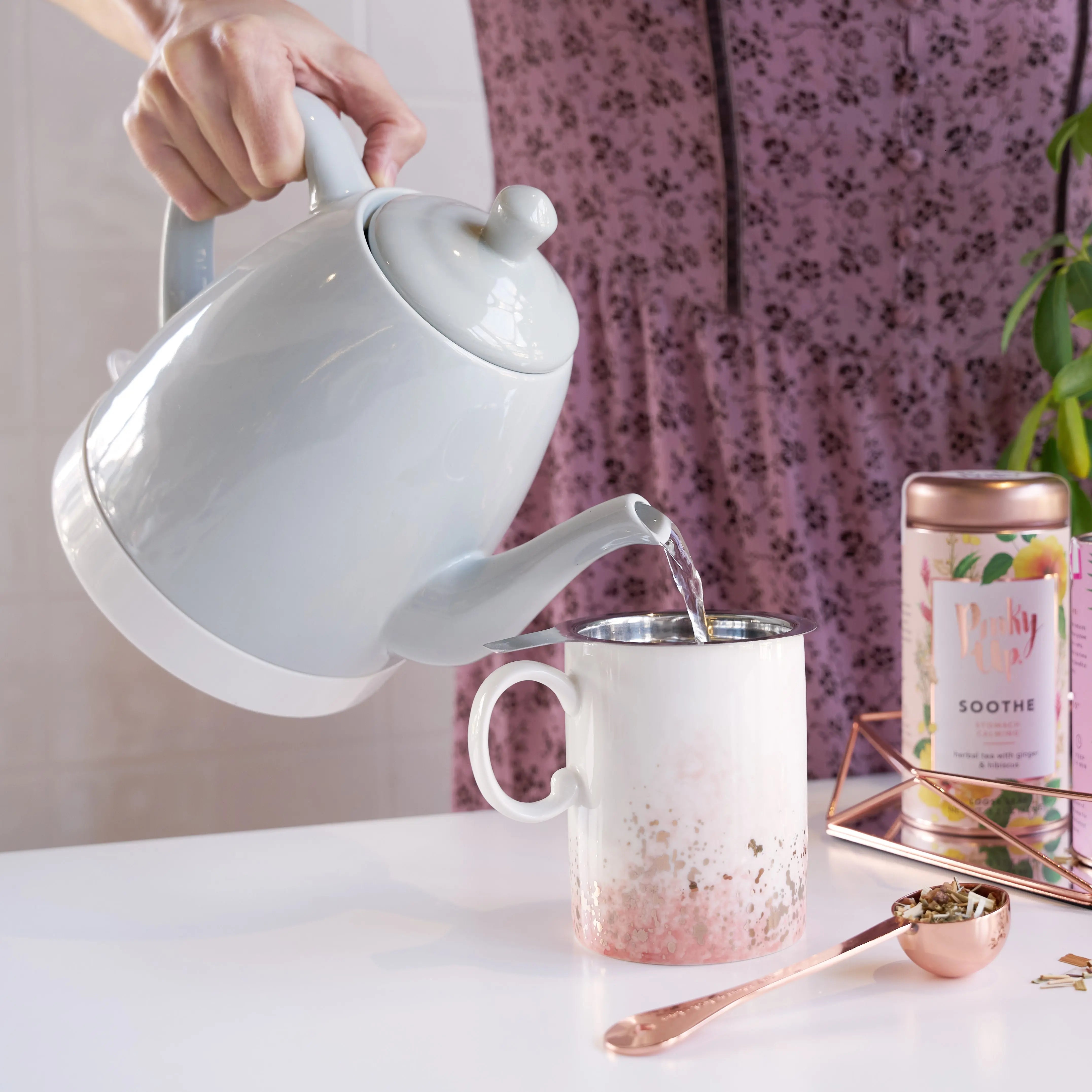 Person pouring tea from a white teapot into a mug on a table with a pink floral curtain in the background.