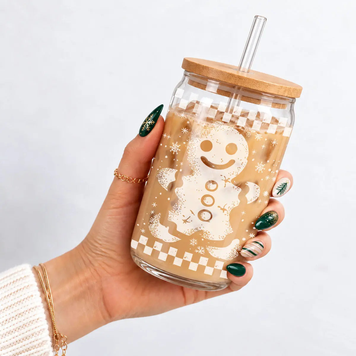 A hand with green and white nail art holds a clear glass jar tumbler with a bamboo lid and clear straw, filled with an iced coffee drink and decorated with a gingerbread man graphic and holiday patterns.