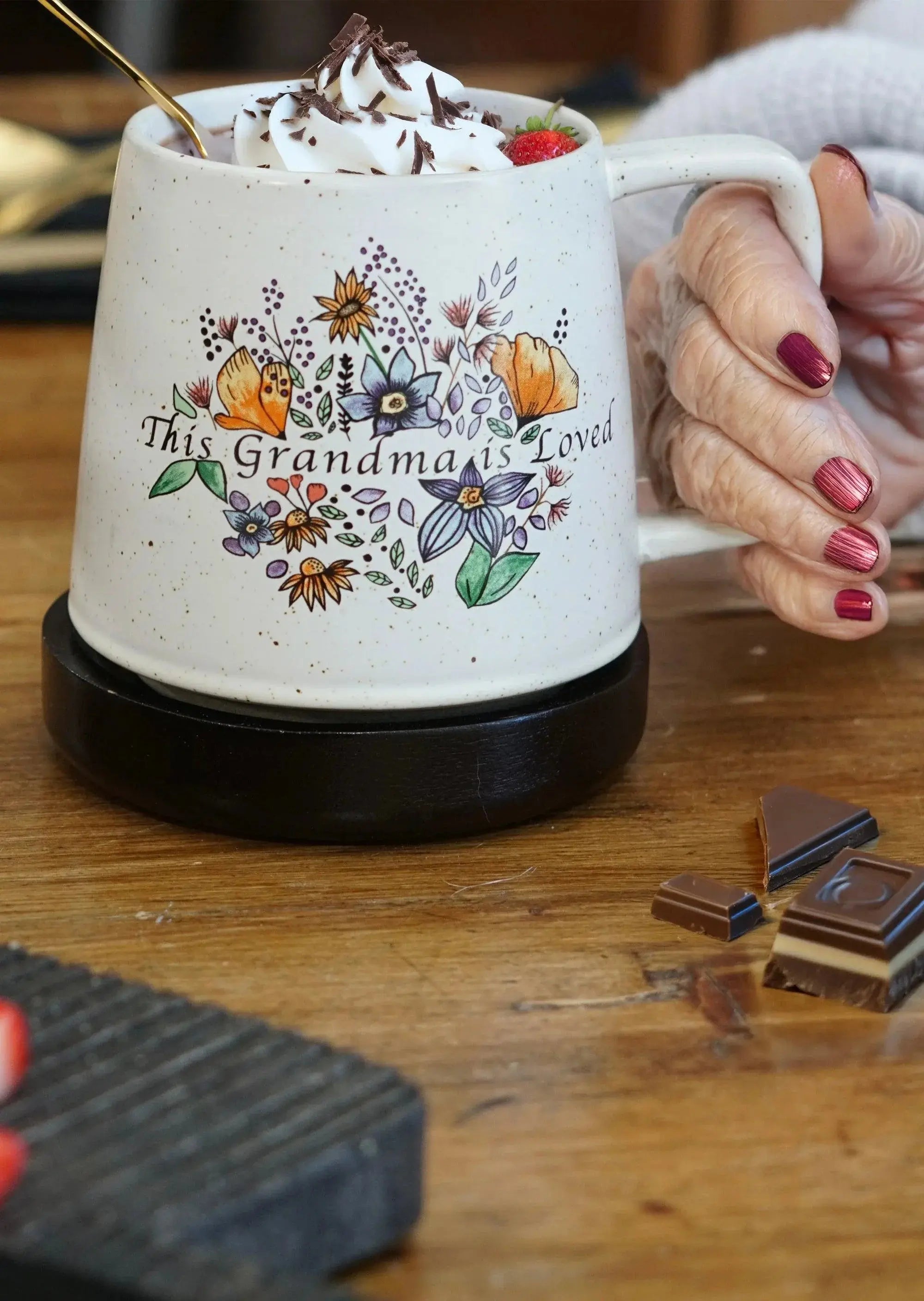 Cozy farmhouse mug with 'This Grandma is Loved' floral design, held by hand, topped with whipped cream, chocolate, and strawberry.