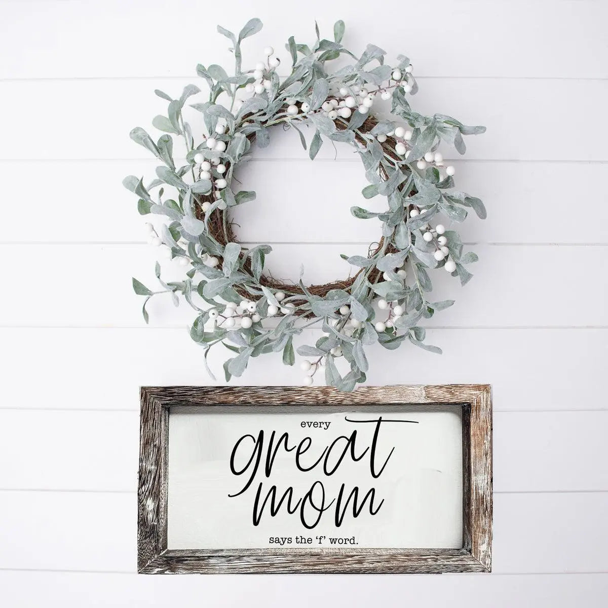 Decorative wreath with greenery and a framed sign on a white wooden background